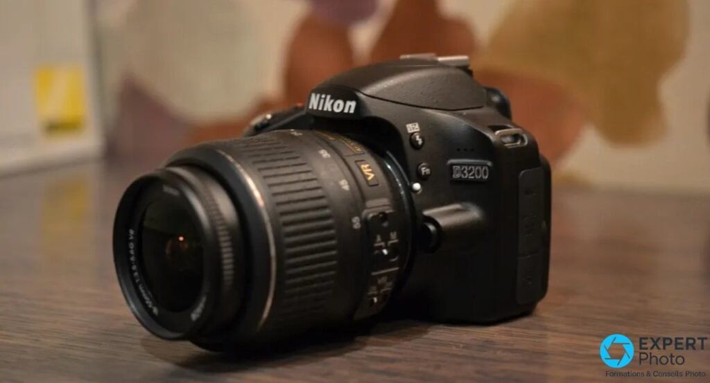 Nikon D3200 noir trois quarts avant droit, objectif Nikkor 18-55mm VR monté, fond aquarelle flou sur table bois