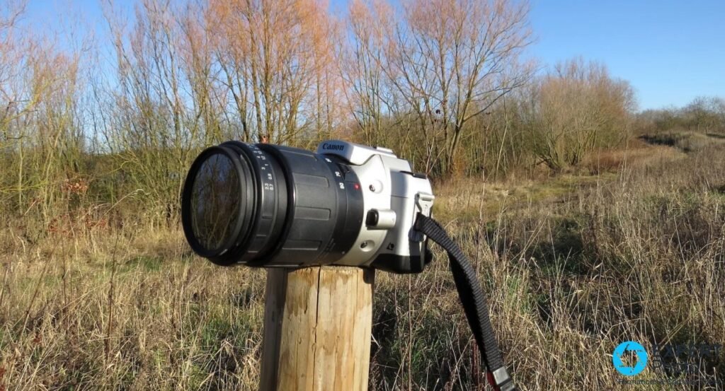 Canon EOS IX7 reflex APS argentique de profil droit, téléobjectif Tamron zoom monté, posé sur poteau bois en nature hivernale 