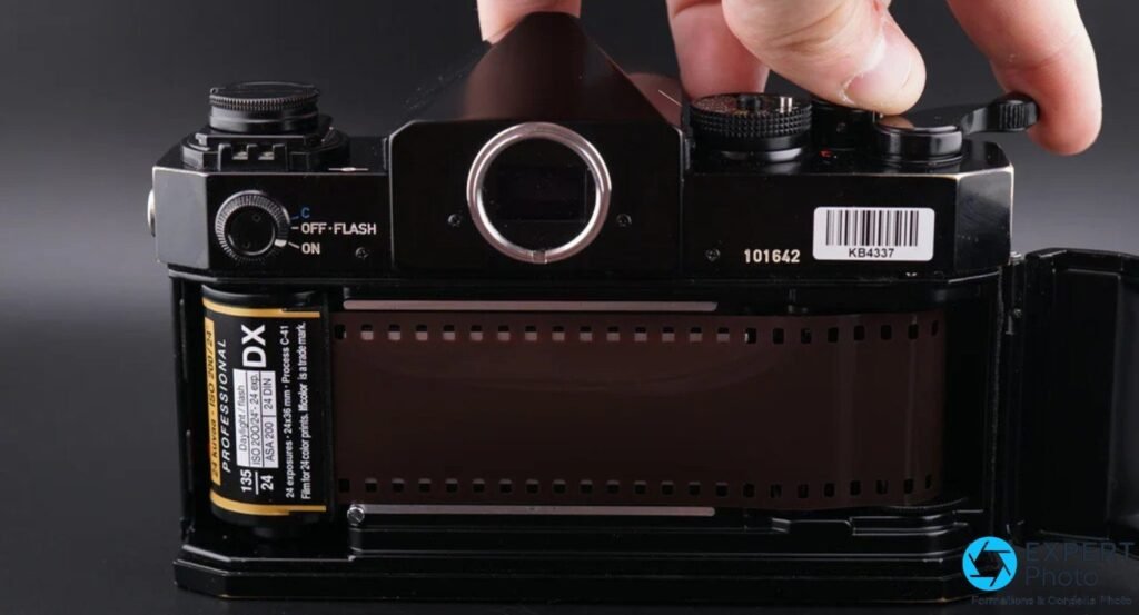 Canon F-1 dos ouvert, pellicule 135 DX ISO 200 chargée côté gauche, bande film tendue, main retirant le pentaprisme du dessus