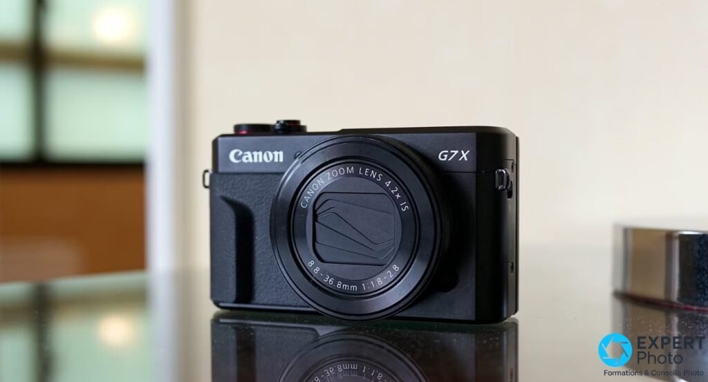 Canon PowerShot G7 X Mark II noir face avant sur table réfléchissante, logos Canon et G7X visibles, fond blanc intérieur