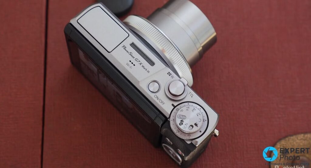 Canon G7X Mark III vu de dos sur surface rouge, écran incliné affichant une scène intérieure, molette et boutons visibles