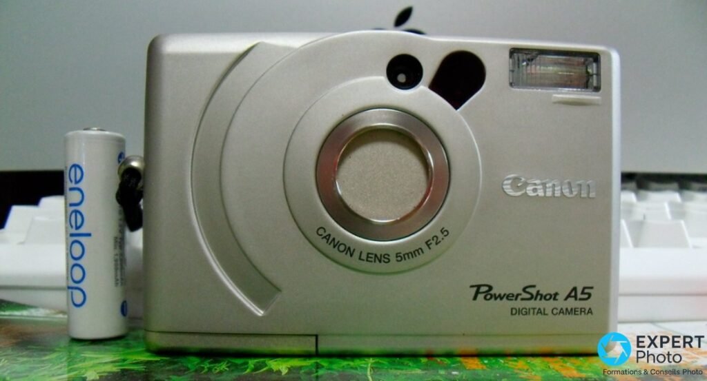 Canon PowerShot A5 vu de face, objectif 5mm F2.5 visible, avec batterie Eneloop AA posée à gauche pour référence de taille