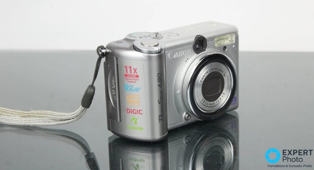 Canon PowerShot A80 argenté, vue trois quarts gauche, zoom 3x et logos DiG!C visibles, fond réfléchissant