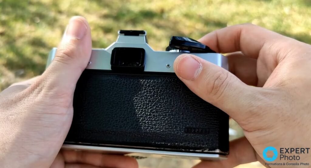 Fujica STX-1N tenu à deux mains, vue de dos : oculaire viseur, dos simili cuir noir, levier armement, fond vert flou
