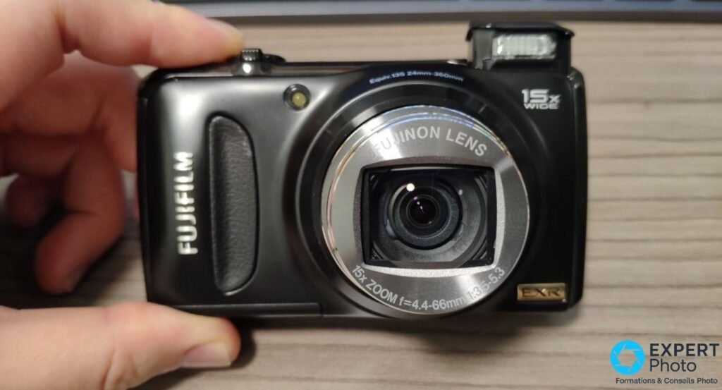 Fujifilm F300EXR tenu en main de face, objectif Fujinon 15x zoom f=4.4-66mm f:3.5-5.3 visible, flash relevé, badge EXR