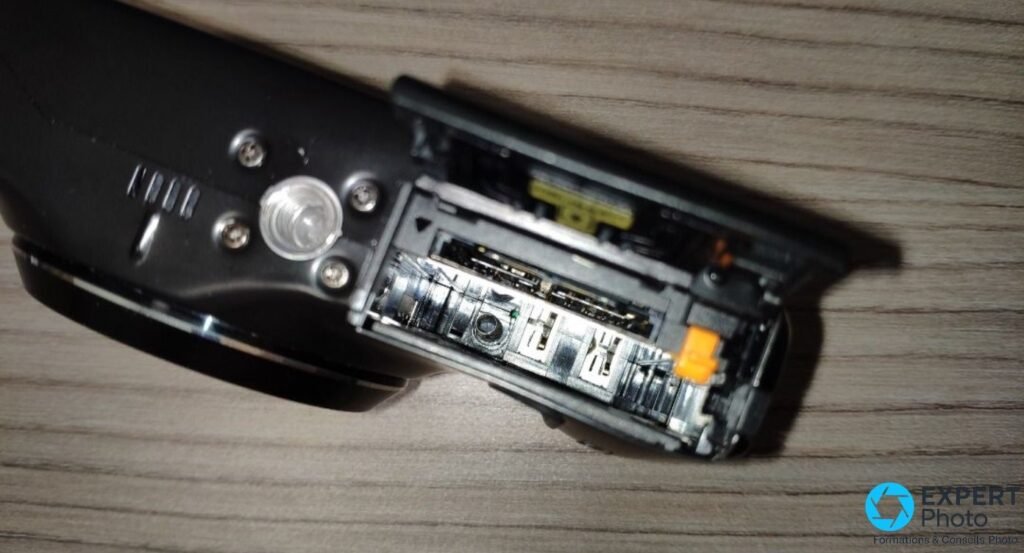 Fujifilm F300EXR vu de dessous, trappe batterie ouverte révélant le compartiment interne et le mécanisme du zoom motorisé
