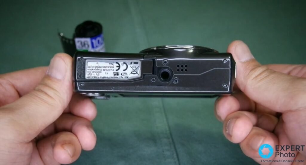 Fujifilm FinePix F200EXR vu de dessous tenu en mains, étiquette constructeur, prise trépied et grille haut-parleur visibles.