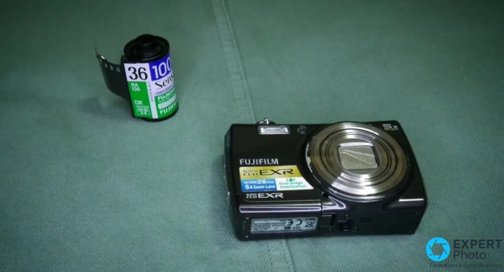 Fujifilm FinePix F200EXR posé sur tissu vert, objectif fermé, aux côtés d'une cartouche de pellicule 35 mm Fujicolor.