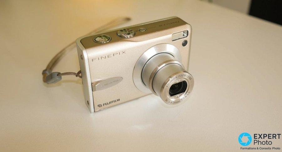 Fujifilm FinePix F30 vu de trois quarts, objectif Fujinon Zoom Lens 3x f=8-24mm f/2.8-5.0 sorti, dragonne grise visible