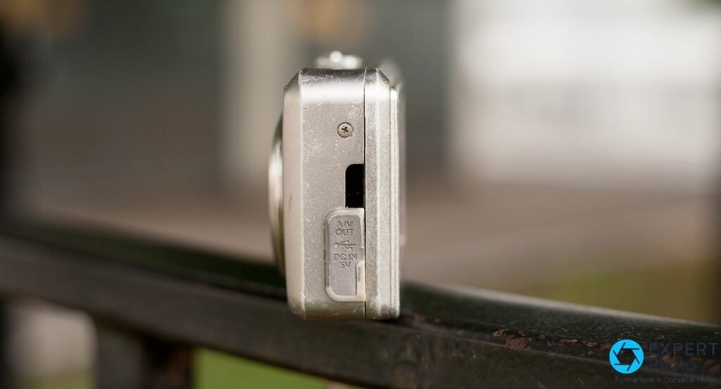 Fujifilm FinePix F31fd tranche gauche posé sur rambarde, trappe ouverte avec ports AV Out, USB et DC In 5V 