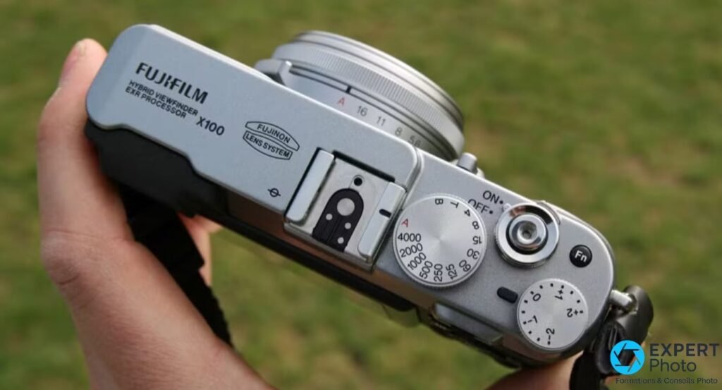 Fujifilm FinePix X100 tenu en main, vue du dessus, molettes vitesse et compensation d'exposition, logo Hybrid Viewfinder 