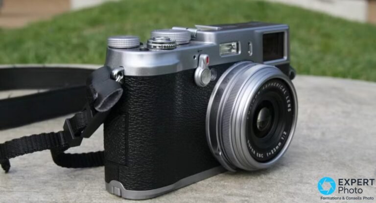 Fujifilm FinePix X100 de trois-quarts sur sol gris, objectif Fujinon 23mm f/2 déployé, sangle noire, fond herbe verte