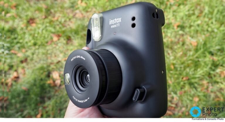 Fujifilm Instax Mini 11 noir tenu en main, objectif tiré en mode selfie, flash visible, fond herbe verte