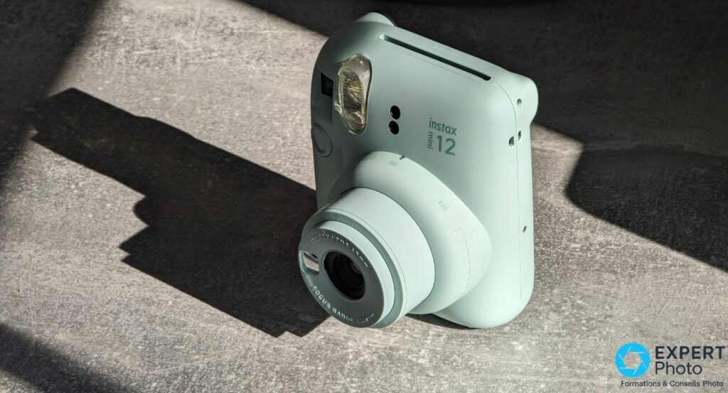 Fujifilm Instax Mini 12 vert menthe en trois quarts avant, objectif déployé et sélecteur ON/OFF visibles, posé sur béton
