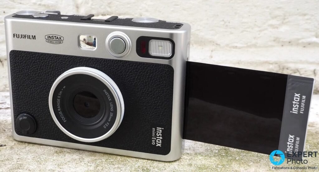 Fujifilm Instax Mini Evo noir et argent, objectif 28mm f/2.0, pack de film instax sorti sur le côté, fond béton