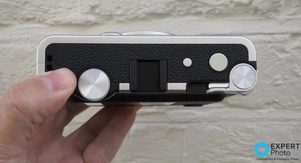 Vue de dessus du Fujifilm Instax Mini Evo tenu en main, deux molettes argentées et levier d'impression central visibles