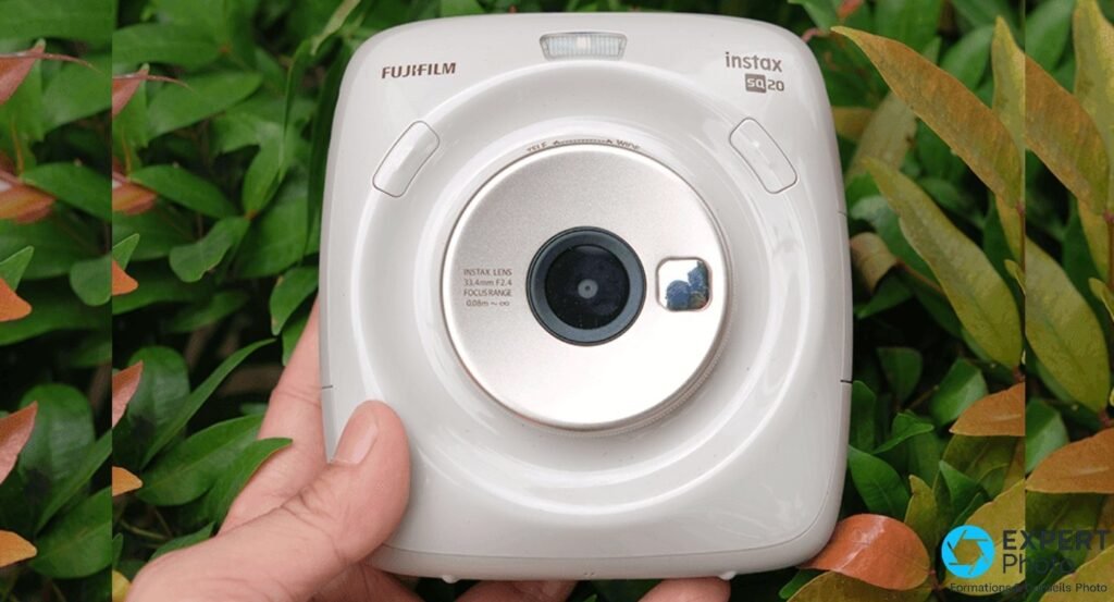 Fujifilm Instax Square SQ20 beige tenu en main, face avant avec objectif 33,4 mm f/2.4 et flash intégré, fond végétal 