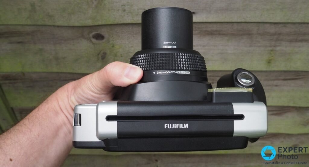 Fujifilm Instax Wide 300 en main, vue de dessus, bague mise au point zones 0,9-3m et 3m-infini, logo Fujifilm visible
