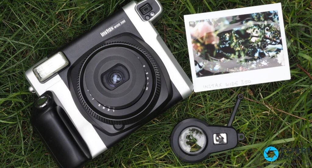 Fujifilm Instax Wide 300 sur herbe verte en flat lay, bonnette gros plan et tirage Wide format paysage avec fleurs à côté