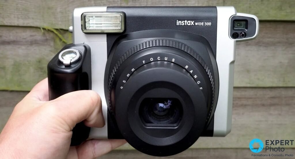 Fujifilm Instax Wide 300 saisi par le grip, face avant, objectif 95mm inscription FOCUS RANGE, flash gauche et viseur droite