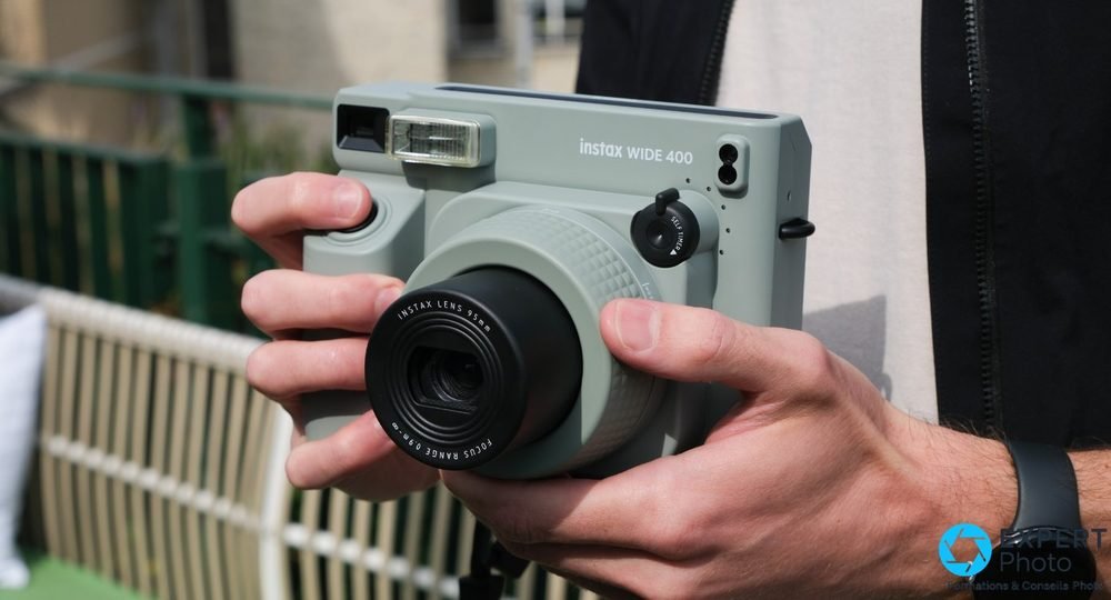 Deux mains tenant l'Instax Wide 400 vert de face en extérieur, objectif 95 mm et flash bien visibles