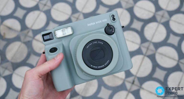 Main tenant l'Instax Wide 400 vert de face, objectif 95 mm, flash et molette retardateur visibles, fond carrelé
