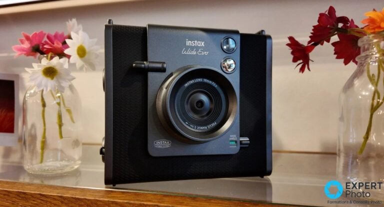 Test Instax Wide Evo : format Wide, créativité et coût à assumer Instax Wide Evo noir posé sur une étagère entre deux vases de fleurs colorées, face avant avec objectif 16mm F2.4