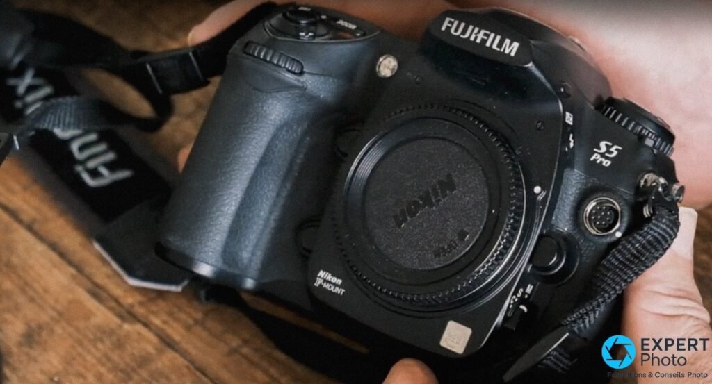 Fujifilm S5 Pro tenu à deux mains, face avant avec bouchon Nikon F-Mount, sangle Fujifilm visible à gauche, fond bois. 