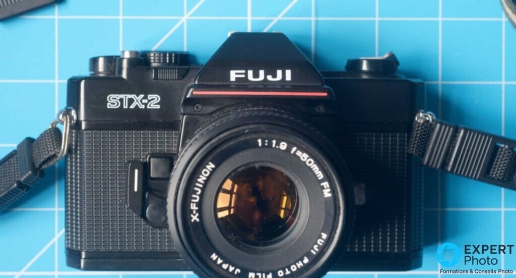  Fujifilm STX-2 noir de face sur tapis de découpe bleu, objectif X-Fujinon 1:1.9 f=50mm, bandoulière noire visible.