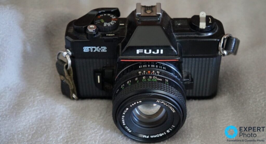 Fujifilm STX-2 vu en trois-quarts avant, objectif X-Fujinon 50mm, bague de mise au point et molette des vitesses visibles.