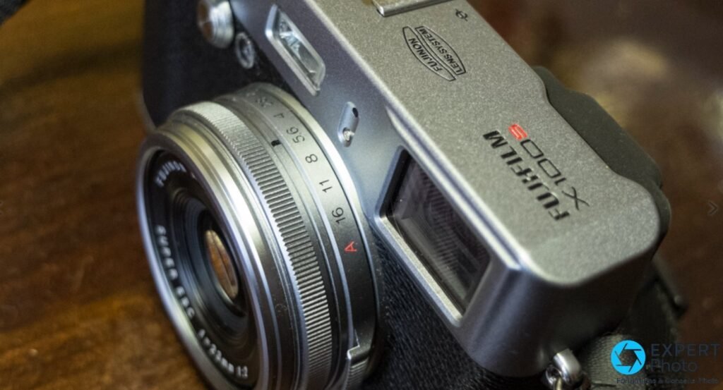 Fujifilm X100S posé en ambiance, boîtier argent et cuir noir, objectif 23 mm Fujinon au centre, masques vénitiens flous.