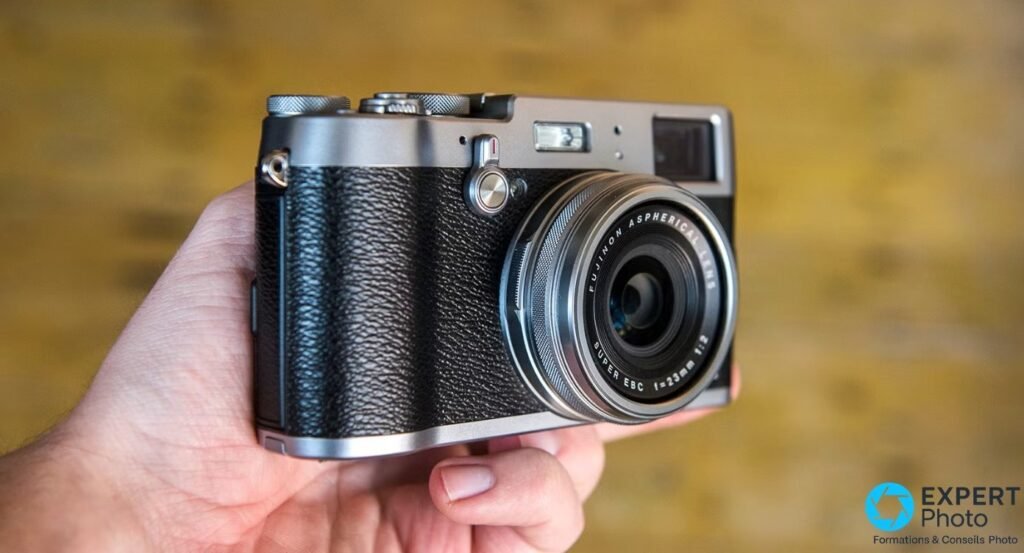 Fujifilm X100T tenu en main, vu de trois quarts avant, objectif Fujinon Aspherical Super EBC 23mm f/2 et flash visibles.