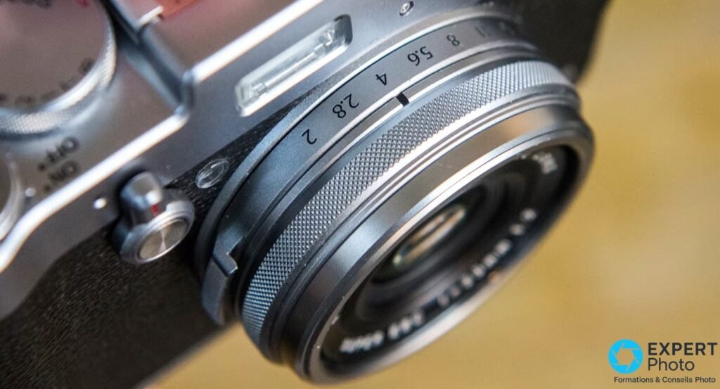 Gros plan sur la bague de diaphragme du Fujifilm X100T, valeurs 2, 2.8, 4, 5.6, 8 gravées, molette vitesses en arrière-plan. 
