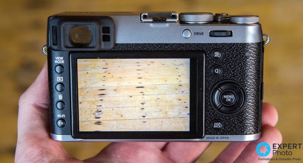 Face arrière du Fujifilm X100T tenu en main, écran LCD 3 pouces non tactile affichant une photo, EVF et boutons visibles.
