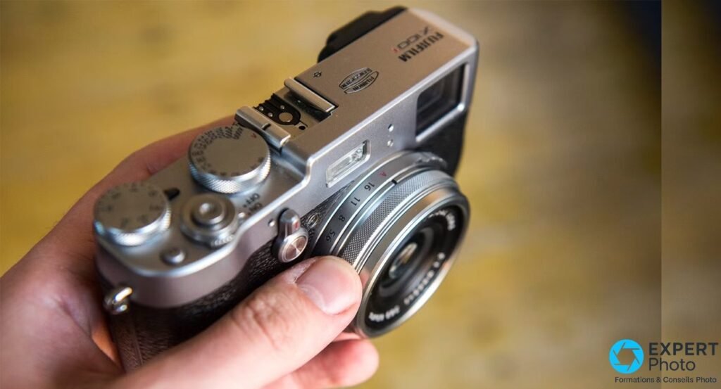 Fujifilm X100T tenu en main, vu en plongée, logo X100T gravé, bague de diaphragme et molette de vitesses visibles.