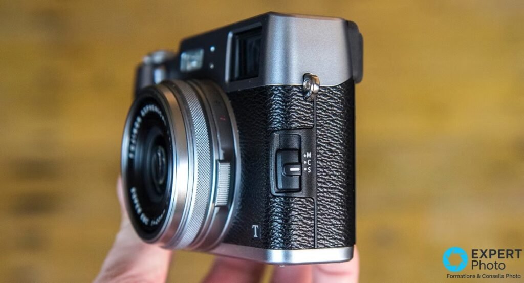 Fujifilm X100T vu de trois quarts droit, tenu en main, sélecteur de mise au point Manuel-Continu-Simple visible en façade.