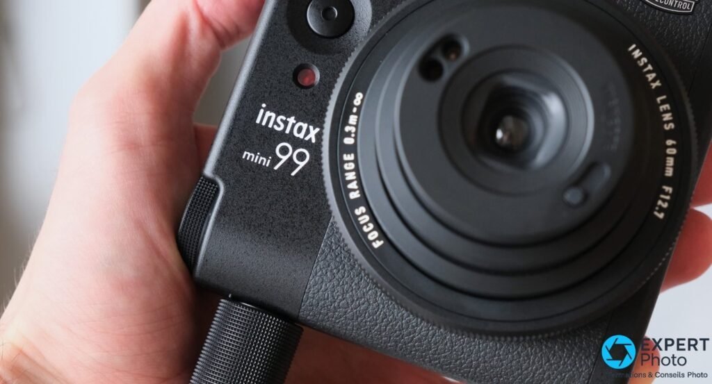 Instax Mini 99 tenu en main, gros plan sur l'objectif 60mm F12.7, grip texturé noir et logo instax mini 99 en blanc
