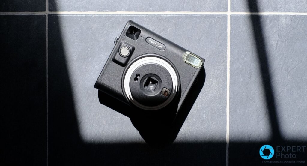 Fujifilm Instax Square SQ40 noir en vue plongeante sur carrelage gris, ombres dures diagonales, flash et viseur visibles