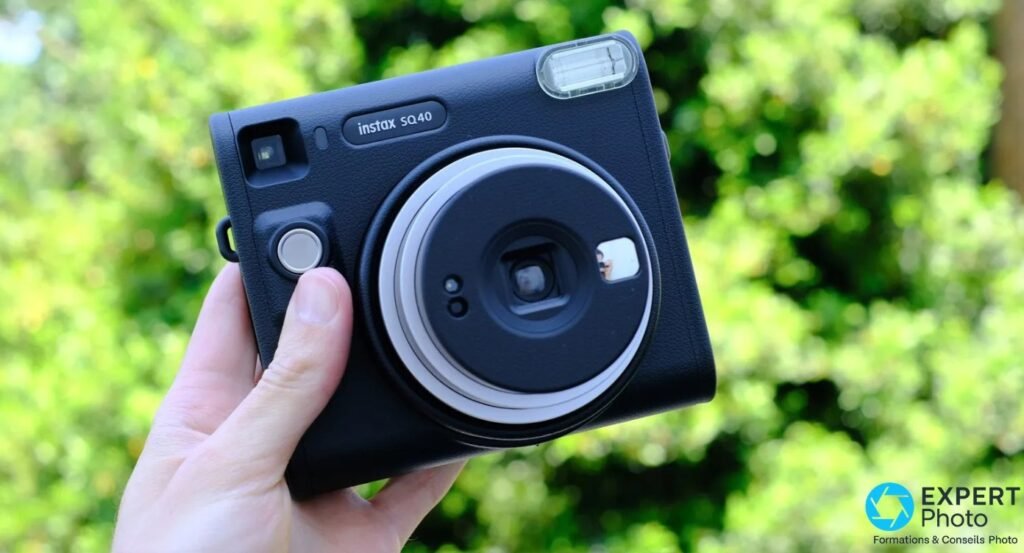 Fujifilm Instax Square SQ40 noir tenu en main à l'extérieur, bokeh vert en arrière-plan, face avant avec déclencheur