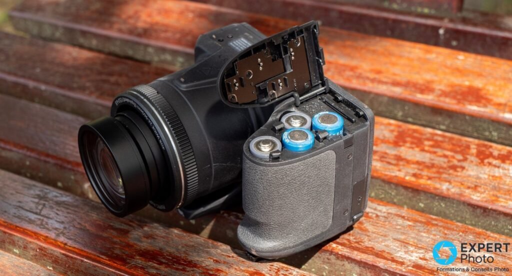 Kodak AZ405 posé sur banc, compartiment piles ouvert révélant 4 piles AA avec embouts bleus NiMH, objectif visible à gauche.