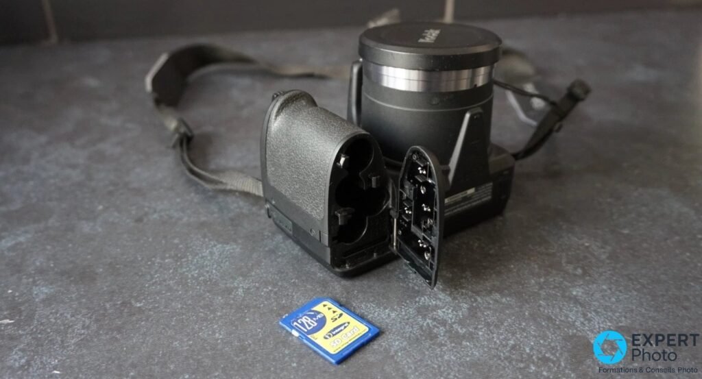 Kodak Pixpro AZ401, trappe inférieure ouverte montrant 4 logements piles AA et slot SD, carte SD 128 Mo posée au premier plan