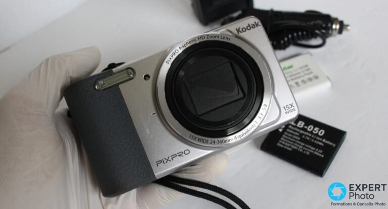 Kodak PIXPRO FZ151 face avant tenu en main gantée, objectif inscrit 15X WIDE 24-360mm, batterie LB-050 et chargeur visibles