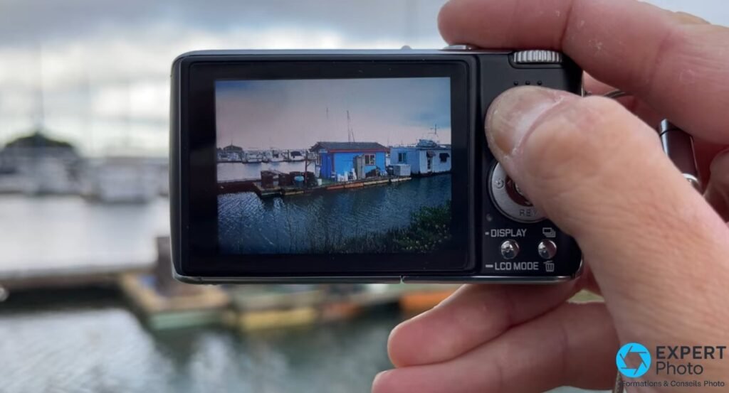 Dos du Leica C-Lux 1 tenu en main, écran 2,5 pouces affichant une photo de port avec maisons flottantes et bateaux