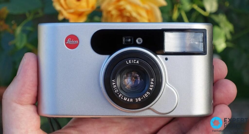 Leica C1 argentique tenu en main devant des roses oranges, façade argent et logo Leica rouge visibles 