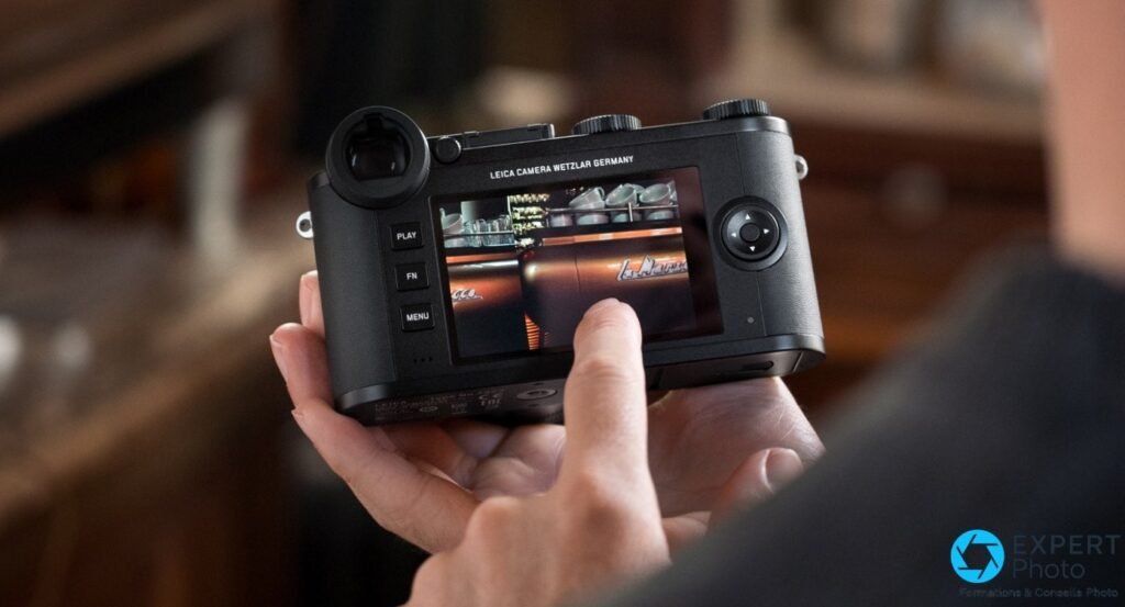 Leica CL vu de dos, mains tenant le boîtier, écran tactile affichant une scène de café, boutons PLAY FN MENU visibles