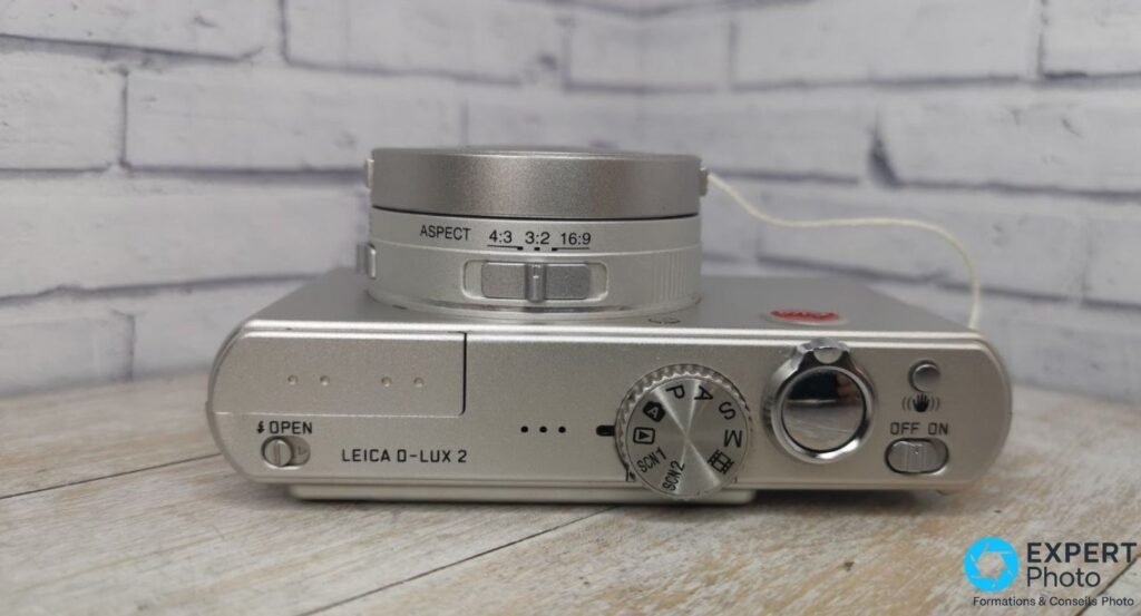 Leica D-Lux 2 vu de dessus, molette de modes P/A/S/M/SCN et commutateur d'aspect 4:3 / 3:2 / 16:9 gravé sur l'objectif
