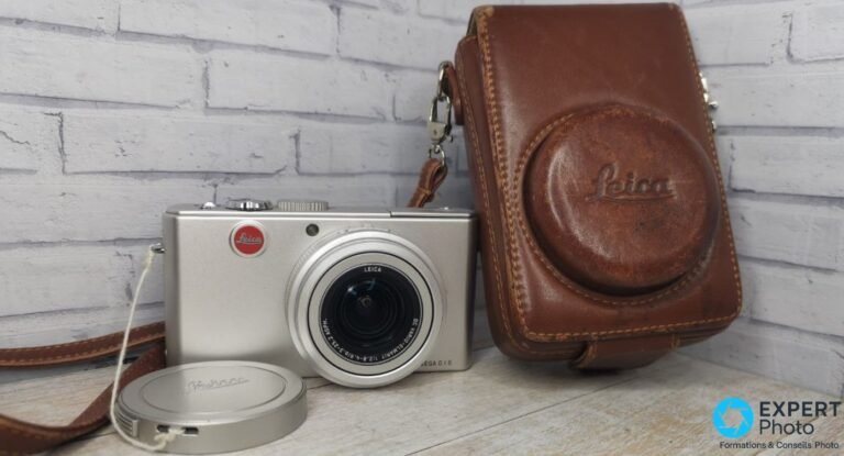 Leica D-Lux 2 argent posé avec son étui en cuir marron Leica, bouchon d'objectif et sangle sur fond de mur de briques blanc