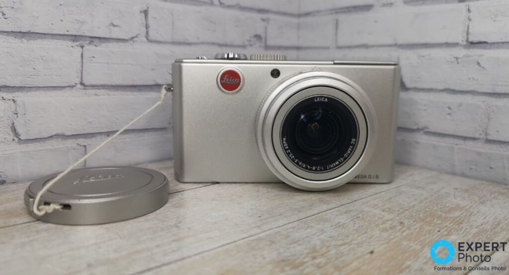 Leica D-Lux 2 argent vu de face trois-quarts, bouchon d'objectif métal détaché relié par cordon, mur de briques blanc en fond