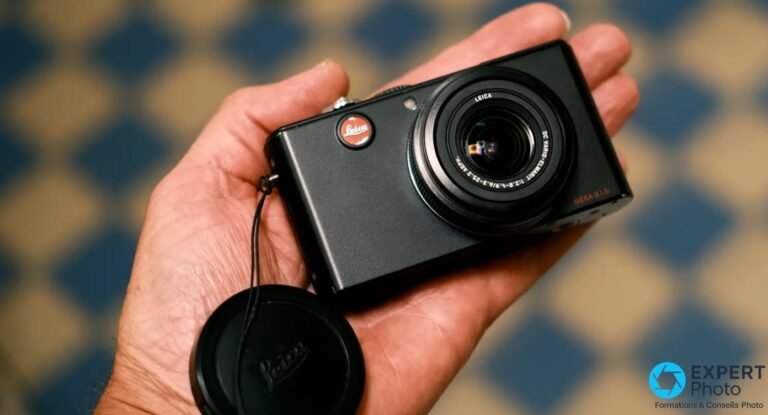 Leica D-Lux 3 noir tenu dans la paume, logo Leica rouge visible, bouchon d'objectif posé au premier plan.