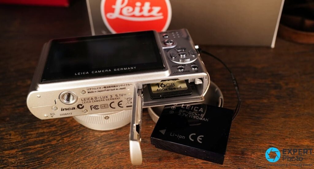 Leica D-Lux 3 argenté, trappe ouverte, carte SD 16 Go et batterie BP-DC4 3,7V posée à côté sur table bois.